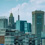 A daytime view of the Baltimore, Maryland skyline featuring a mix of modern and historic skyscrapers, including the iconic gold-topped 10 Light Street building, under a blue sky with light clouds. Waterfront residential buildings with balconies are visible in the foreground.
