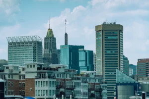 A daytime view of the Baltimore, Maryland skyline featuring a mix of modern and historic skyscrapers, including the iconic gold-topped 10 Light Street building, under a blue sky with light clouds. Waterfront residential buildings with balconies are visible in the foreground.