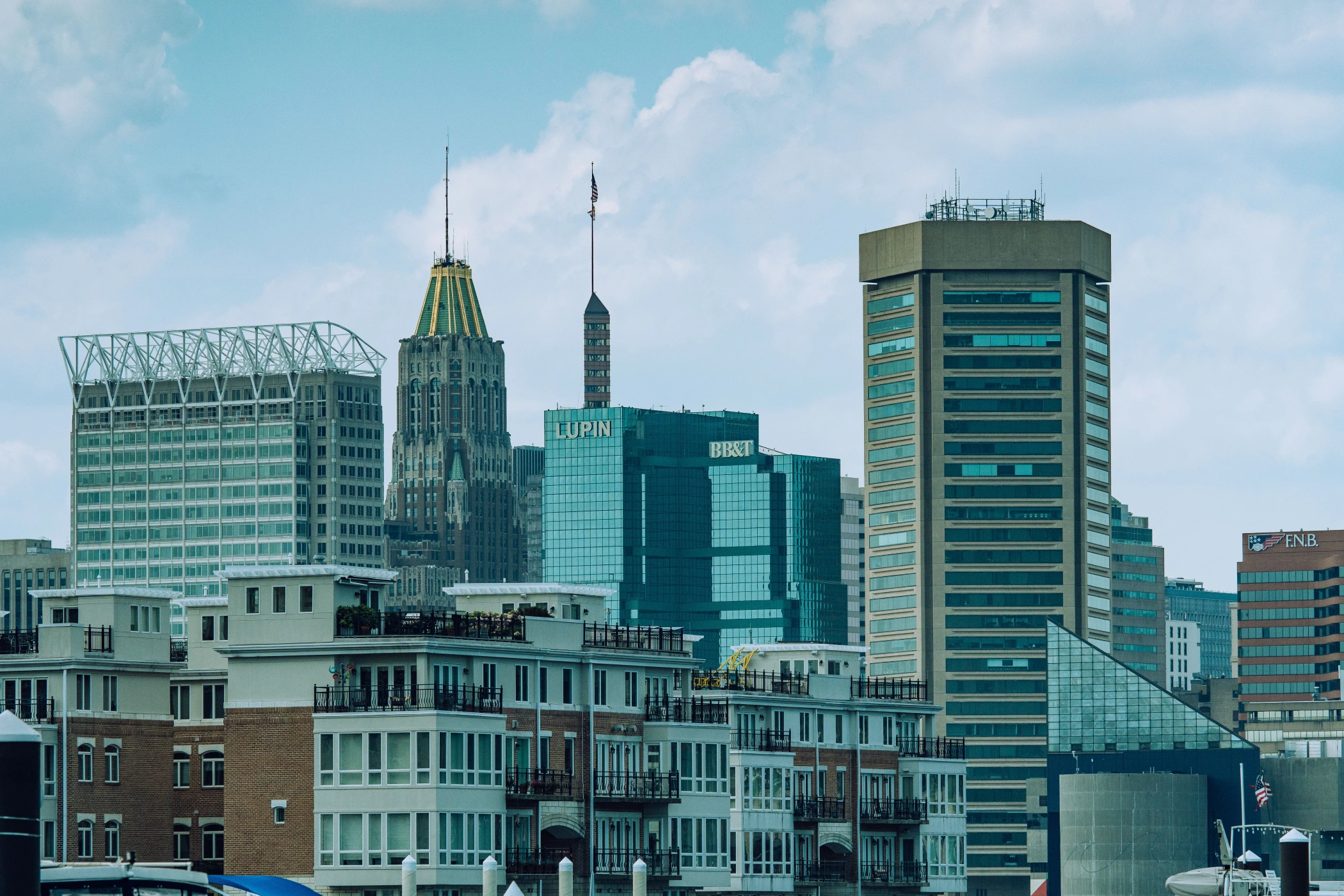 A daytime view of the Baltimore, Maryland skyline featuring a mix of modern and historic skyscrapers, including the iconic gold-topped 10 Light Street building, under a blue sky with light clouds. Waterfront residential buildings with balconies are visible in the foreground.