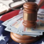A close-up, high-angle photograph depicting powerful symbols of the American legal system and financial law. In the center, a polished wooden judge's gavel stands upright on its circular sound block, holding down a fanned-out stack of one-hundred-dollar US bills. The entire arrangement rests on a draped American flag, with its white stars against a blue background visible in the lower left. In the softly blurred background, a pair of metallic handcuffs and an open spiral-bound notebook are partially visible, evoking themes of justice, bail, court settlements, or financial crime.