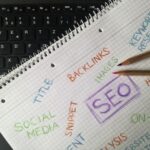 A top-down shot of a wire-bound notebook with grid paper laid over a black computer keyboard. The notebook page contains hand-drawn marketing notes written in colored pencils, including the words "SEO" boxed in purple, "BACKLINKS" in orange, "SOCIAL MEDIA" and "IMAGES" in green, and "KEYWORD RESEARCH" in blue. Two wooden colored pencils rest on the right side of the page