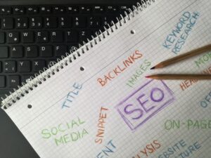 A top-down shot of a wire-bound notebook with grid paper laid over a black computer keyboard. The notebook page contains hand-drawn marketing notes written in colored pencils, including the words "SEO" boxed in purple, "BACKLINKS" in orange, "SOCIAL MEDIA" and "IMAGES" in green, and "KEYWORD RESEARCH" in blue. Two wooden colored pencils rest on the right side of the page