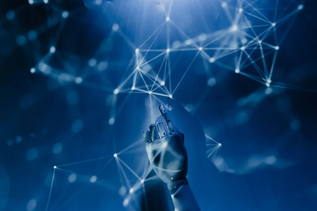 A conceptual digital image showing a white robotic hand reaching upward, its index finger pointing toward a complex constellation of glowing white nodes and intersecting data lines. The entire scene is bathed in deep shades of blue, creating a high-tech, futuristic neural network aesthetic.