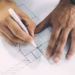 Hands using a ruler and pen to draft a compliant retail dispensary floor plan layout on architectural blueprints.