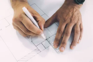 Hands using a ruler and pen to draft a compliant retail dispensary floor plan layout on architectural blueprints.