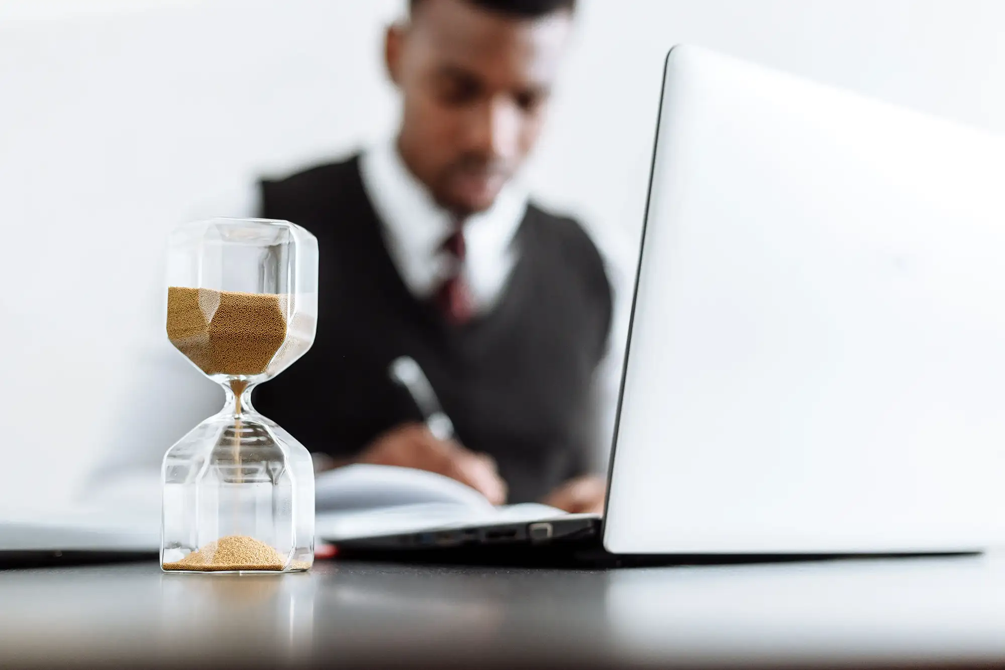 An hourglass with sand running through it in front of a business professional working on a laptop and taking notes.
