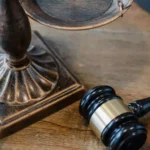 Bronze scales of justice resting next to a wooden gavel and a legal binder on a wooden desk.