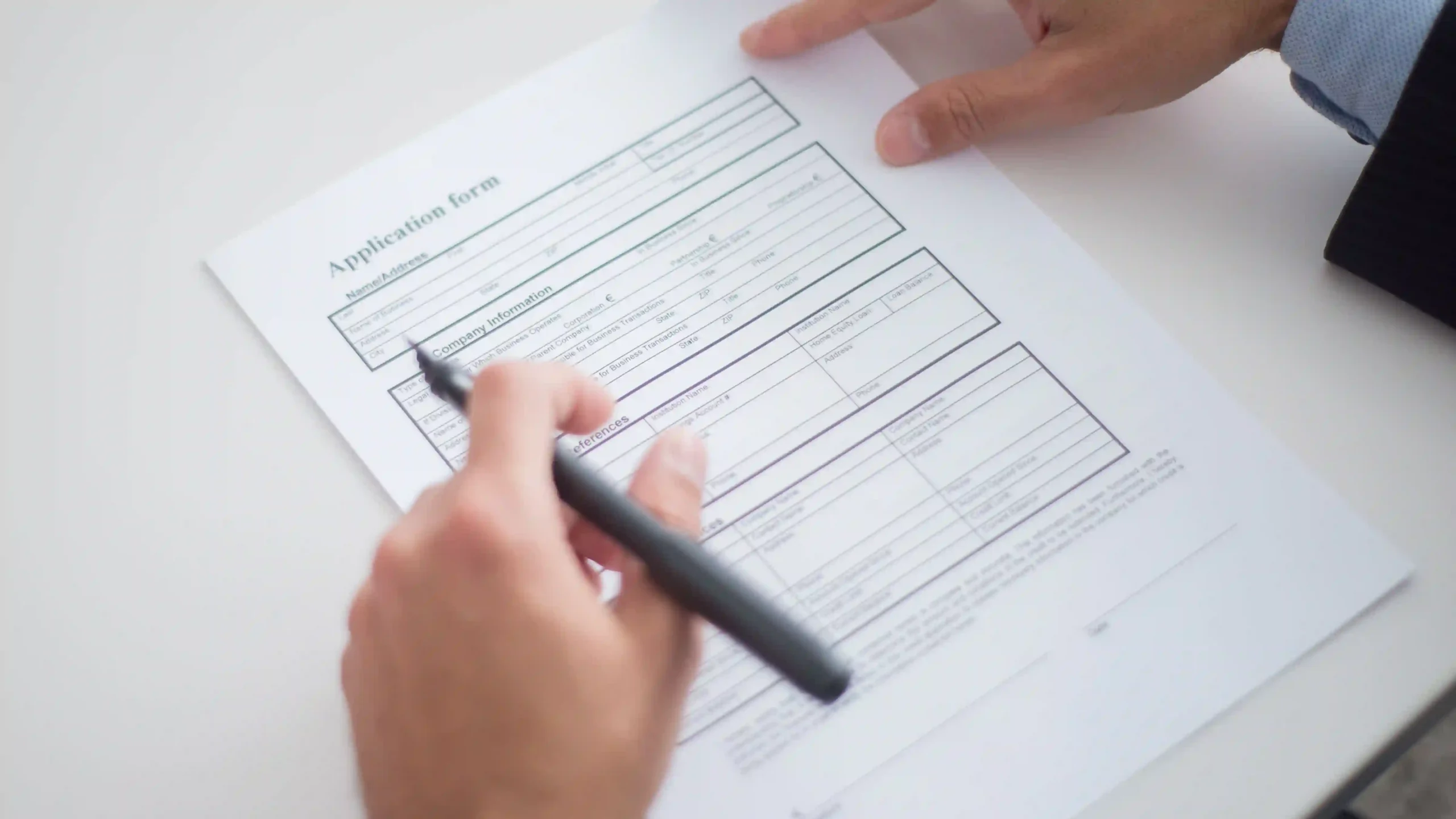 Close-up of a person in a business suit holding a pen while filling out an application form on a white desk.