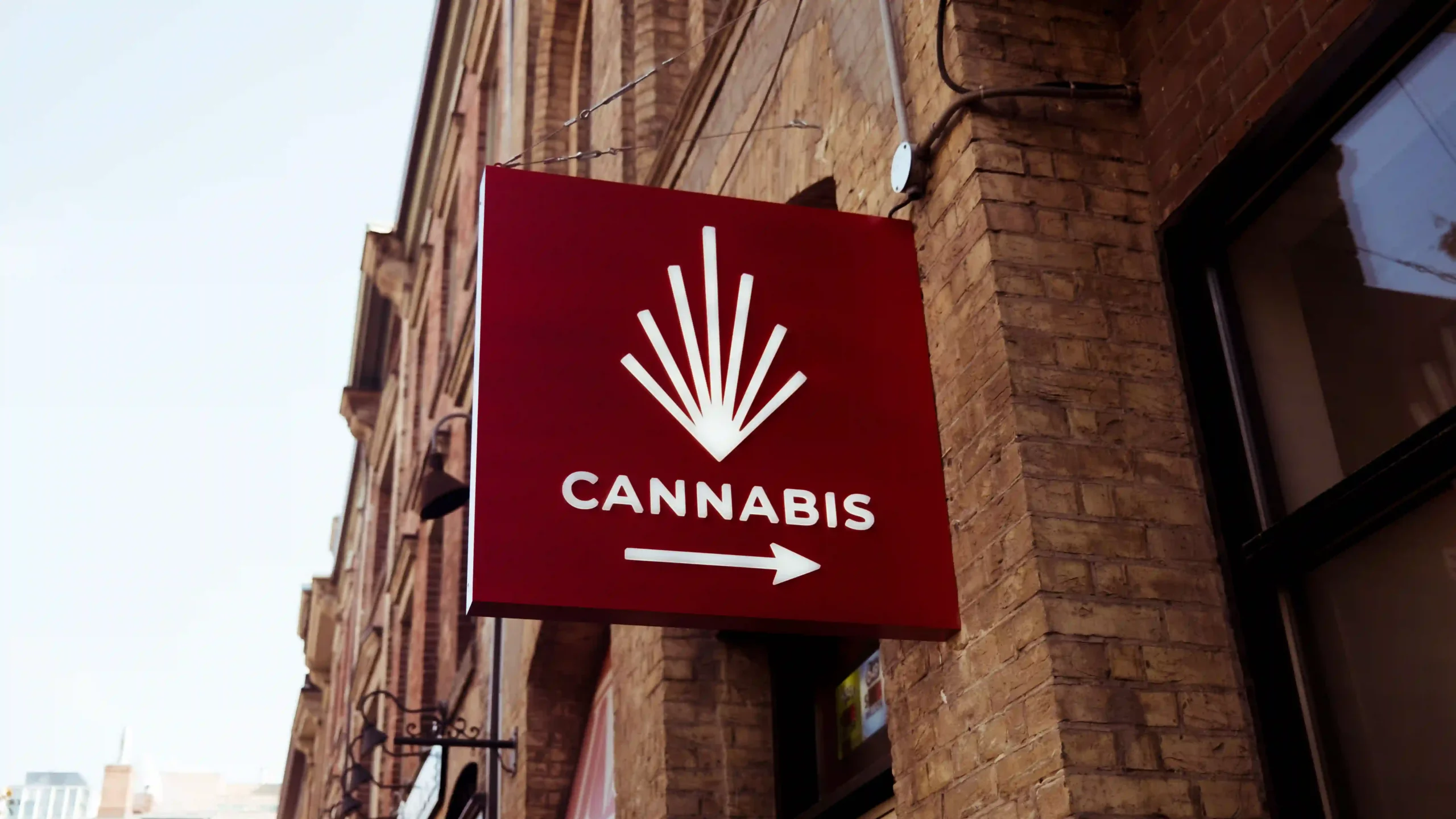 A red outdoor sign hanging on a brick building with a stylized leaf graphic, the word CANNABIS, and an arrow pointing to the right